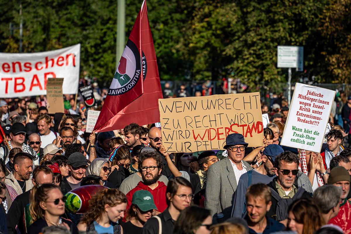 Das Foto von © Björn Obmann zeigt einen Ausschnitt aus der Demo. Sichtbar sind eine Fahne der "Antikolonialen Aktion" eine Papptafel mit dem Text "Wenn Unrecht zu Recht wird, wird Widerstand zur Pflicht" sowie eine Tafel mit "Jüdische Mitbüger:innen wegen Israelkritik unter Extremismusverdacht?!? Wann hört die Freak-Show endlich auf?". Halb verdeckt ist ein Transparent mit dem Text: "Die Würde des Menschen ist antastbar" zu sehen.