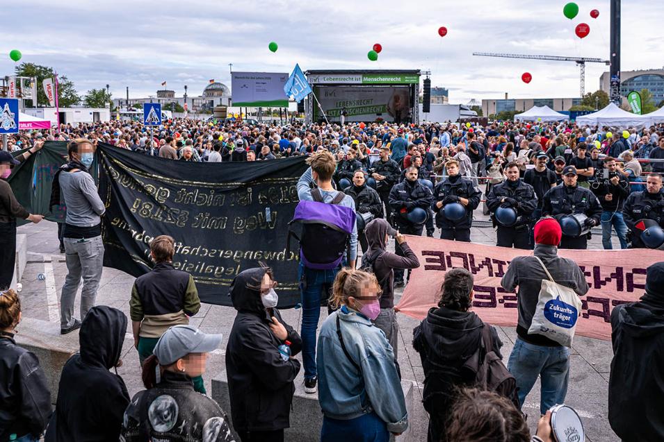 Farbfotografie einer Demonstration auf dem Platz der Republik vor dem Reichstagsgebäude in Berlin. Im Hintergrund versammelt sich eine große Menschenmenge vor einer Bühne mit der Aufschrift „Lebensrecht ist Menschenrecht“. Über der Menge schweben rote und grüne Luftballons. Im Vordergrund stehen Demonstrierende mit schwarzen und rosafarbenen Transparenten einer Gegenprotestaktion. Ihnen gegenüber hat sich eine Reihe von Polizeibeamten in schwarzer Einsatzkleidung mit Schutzhelmen aufgestellt. Die Aufnahme zeigt das Aufeinandertreffen einer Demonstration gegen Schwangerschaftsabbrüche und ihres Gegenprotests.
