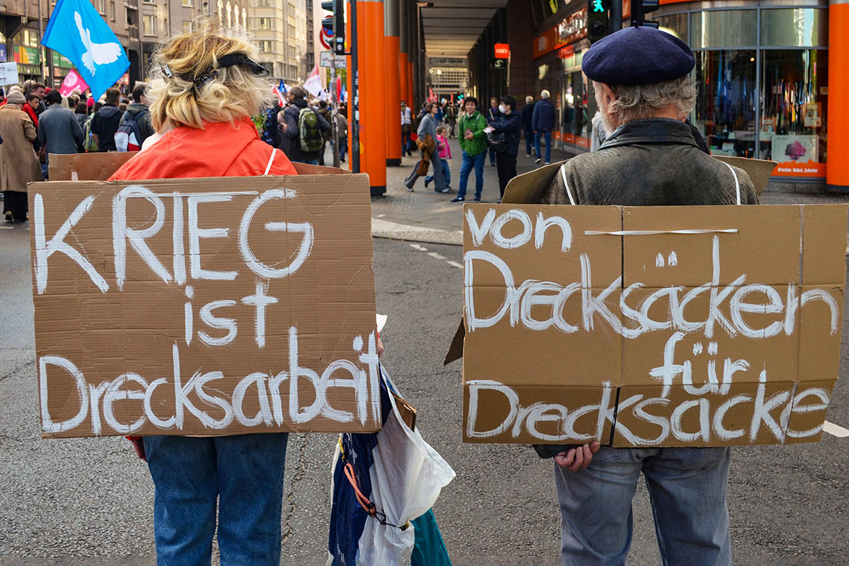 Das Foto von © Monika von Wegerer zeigt zwei Menschen von hinten, die sich zwei ergänzende Texttafeln umgehängt haben: Die links: "Krieg ist Drecksarbeit" die rechts: "von Drecksäcken für Drecksäcke"