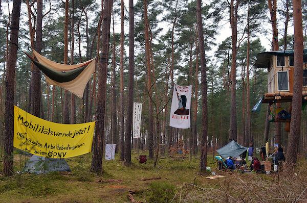 Foto: © Monika v. Wegerer via Umbruch Bildarchiv Das Foto von Monika v. Wegerer zeigt ein Waldstück. Rechts ein Baumhaus, unter dem einige Personen sitzen. In der Mitte des Bildes ist ein Transparent mit Pauli, dem Maulwurf und der Aufschrift "Wuhli bleibt!" zu sehen, links im Bild ist ein Transparent mit der Forderung "Mobilitätswende jetzt! Nur mit guten Arbeitsbedingungen im ÖPNV!" zu sehen. Darüber hängt ein Schlafsack zwischen zwei Bäumen.