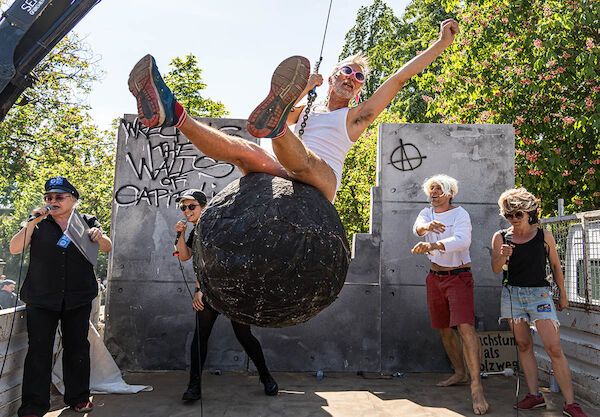 Das Foto von © Kinkalitzken zeigt eine Einlage mit 5 Personen auf einer Baustelle. Auf der Betonwand steht: "Wreck the Walls of Capitalism" und ein Anarchiesymbol. Eine Person schaukelt auf einer Abrißbirne, ein als Vertreter der Spezial-Enteignungs-Kräfte (S.E.K.) Grunewald verkleideter Mensch spricht in ein Mikrofon