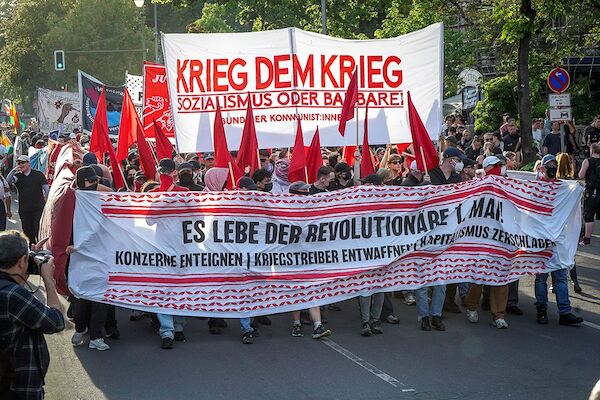 Das Foto zeigt ein Fronttransparent mit dem Text: "Es lebe der evolutionäre 1. Mai! - Konzerne enteignen - Kriegstreiber entwaffnen - Kapitalismus zerschlagen" Dahinter ein Hochtransparen mit dem Text "Krieg dem Krieg - Sozialismus oder Barbarei" dazwischen lauffen teils vermummte Personen mit roten Fahnen