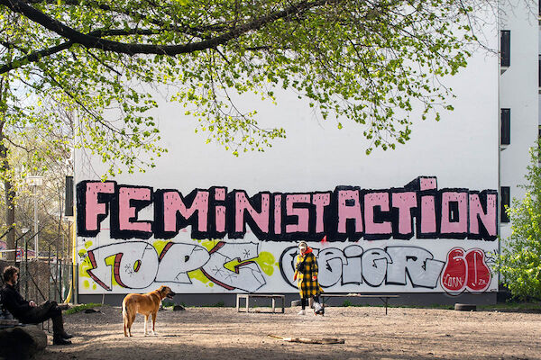 Das Foto zeigt ein Grafitty auf einer Mauer in einem Park. Der Text: "Feminist Action". Davor steht eine Person mit einem Stöckchen in der Hand, ein Hund sieht zu ihr. Eine weitere Person sitzt auf einer Bank und schaut beiden zu.