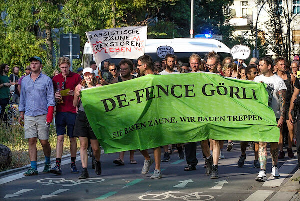 Foto: © heba / Umbruch Bildarchiv Das Foto von © heba zeigt Demonstrant:Innen mit einem grünen Transparent, auf dem der Text "DE-FENCE GÖRLI - Sie bauen Zäune, wir bauen Treppen" steht. Dahinterist auch ein Hichtransparent mit dem Text "Rassistische Zäune zerstören Leben" zu sehen.