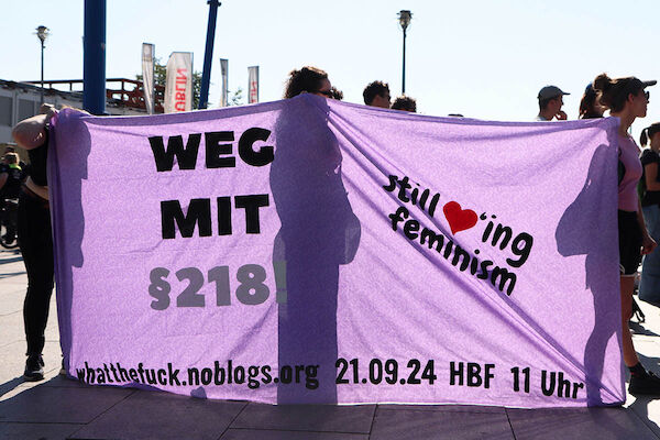 Das Foto zeigt ein Transparent, durch das die Sonne scheint. Dahinter stehen 3 Personen, die es als Seitentransparent halten. Der Text auf dem Transpi: "Weg mit §218, still loving feminism sowie whatthefuck.noblogs.org 21.09.24 HBF 11 Uhr"