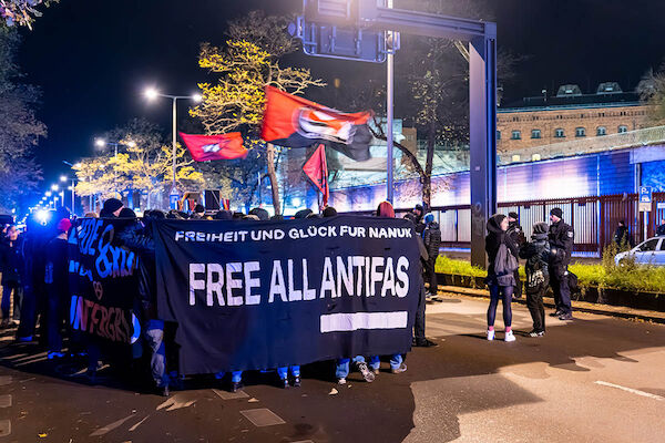 Das Foto zeigt die Demo vor der JVA Moabit. Zu sehen ist das Fronttransparent mit dem Text "Freiheit und Glück für Nanuk - Free all Antifas" sowie einige Seitentransparente und Teilnehmer:*Innen der Demo, die nach rechts Richtung JVA blicken