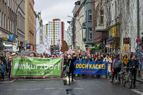 Das Foto zeigt die Demo mit 2 Fronttransparenten: "Wir sind alle #unkürzbar! Ein Berlin für alle" und "Is doch Kacke! #Meckerchor". Dahinter diverse weitere Transparente und Texttafeln