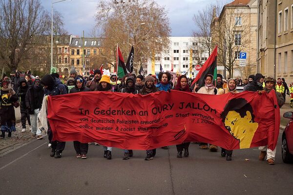 Das Foto zeigt das Fronttransparent mit dem Text "In Gedenken an Oury Jalloh - von deutschen Polizisten ermordet - vom Staat vertuscht, 7.1.2005 Police murder & racist state cover-up" neben einem Bild von Oury Jalloh