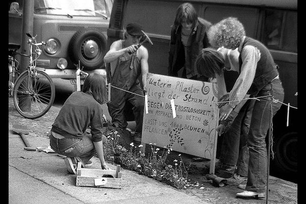 Ein paar Menschen stellen ein Plakat auf. Der Text: "Unter dem Pflaster liegt der Strand - gegen die Trostlosigkeit von Asphalt und Beton - lasst uns 1000 Blumen pflanzen". Foto: © Monika von Wegerer | Schöneberg, Pallasstr., Aktion "unter dem Pflaster liegt der Strand," 1979