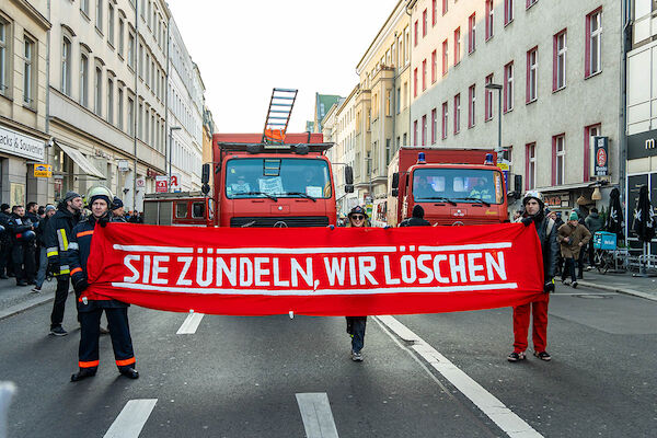 Das Foto zeigt das von Menschen in Feuerwehrkluft getragene rote Transparent mit der Aufschrift: "Sie zündeln - wir löschen" Dahinter sind einige Feuerwahrfahrzeuge und weitere Demonstrant:*Innen zu sehen