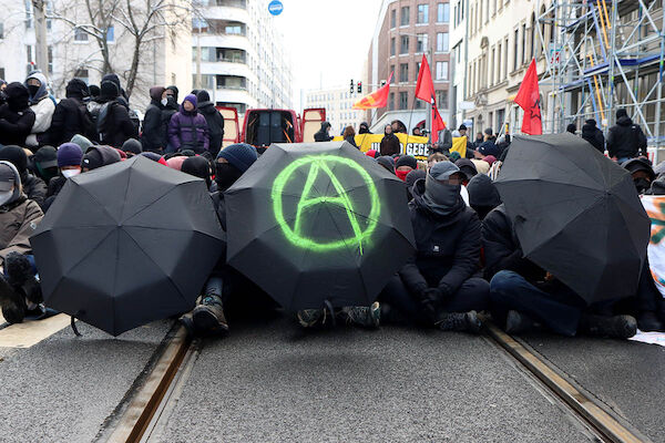 Das Foto von © Kinkalitzken zeigt eine Sitzblockade, verborgen hinter drei schwarzen Regenschirmen, der mittlere bemalt mit dem Anarchiezeichen Ⓐ Dahinter stehen einige Menschen.