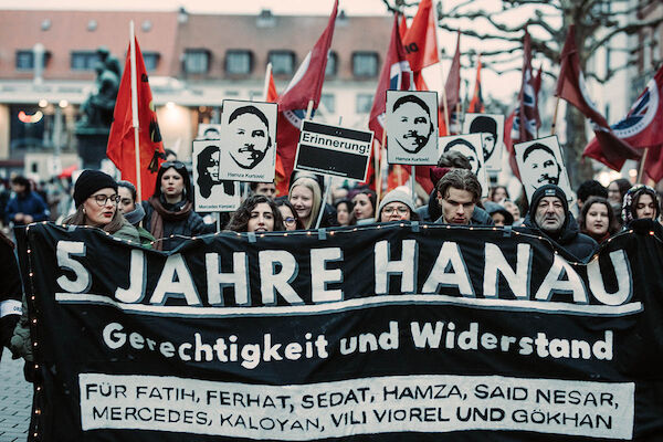 Das Foto zeigt das Transparent mit dem Text: "5 Jahre Hanau - Gerechtigkeit und Widerstand - Für Ferhat Unvar, Hamza Kurtović, Said Nesar Hashemi, Vili Viorel Păun, Mercedes Kierpacz, Kaloyan Velkov, Fatih Saraçoğlu, Sedat Gürbüz und Gökhan Gültekin". Dahinter zahlreiche Menschen mit Fahnen und Tafeln mit den Gesichtern und Namen der Opfer.