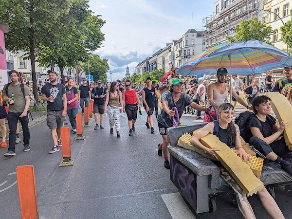 Das Foto von © Sabine Scheffer zeigt einen Ausschnitt der Demo. Im Vordergrund sitzen Menschen auf einem fahrbaren Sofa unter einem Sonnenschirm,