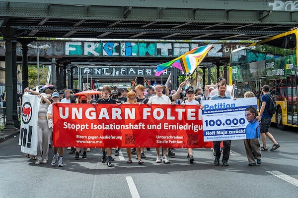 Das Foto zeigt die Demo mit dem Fronttransparent mit dem Text "Ungarn foltert! - Stoppt Isolationshaft und Erniedrigung" sowie jeweils einem Barcode auf die "Eltern gegen Auslieferung" ww.kanu.de und zur Petition. Daneben läuft eine Person mit einem Plakat auf der neben einem Foto von Maja nähere Informationen zur Petition, unter anderem 100.000 Unterzeichner:Innen vermerkt sind.