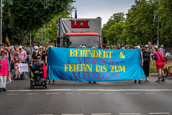 Das Foto von © Björn Obmann zeigt das Fronttransparent mit der Aufschrift "Behindert & verrückt - Feien bis zum Umfallen!" Dahinter fährt ein Lautsprecherwagen. Drumherum viele Menschen unter anderem einige in Rollis.