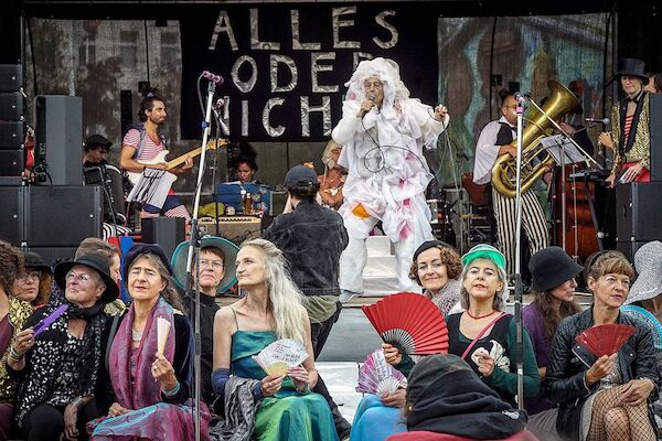 Das Foto zeigt einen Ausschnitt der Bühne mit der Band im Hintergrund vor dem Transparent mit dem Text "Alles oder nichts". Auf der Bühne singt eine mit einem wallenden weißen Ganzkörperkostüm verkleidete Person während im Vordergrund ein Dutzend kostümierte weitere Mitglieder des Ensembles sitzen.