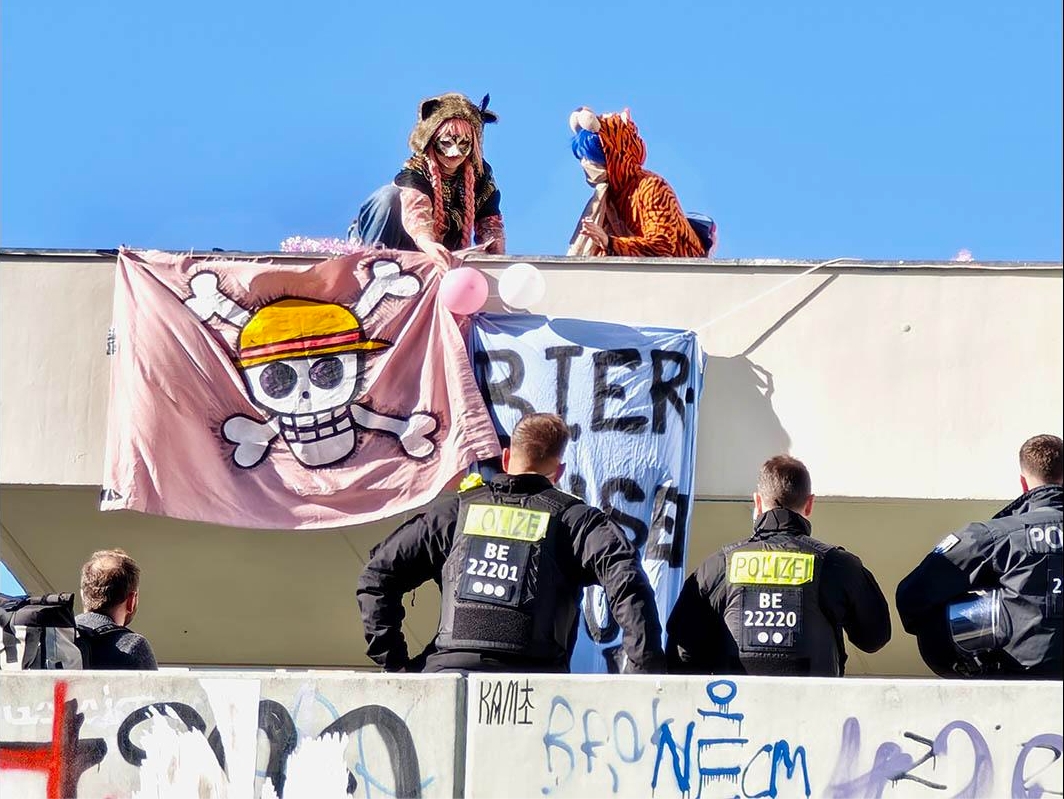 Das Foto von © Sabine Scheffer zeigt kostümierte Aktivist:Innen beim Aufhängen von Transparenten, die dabei von Bullen beobachtet werden.