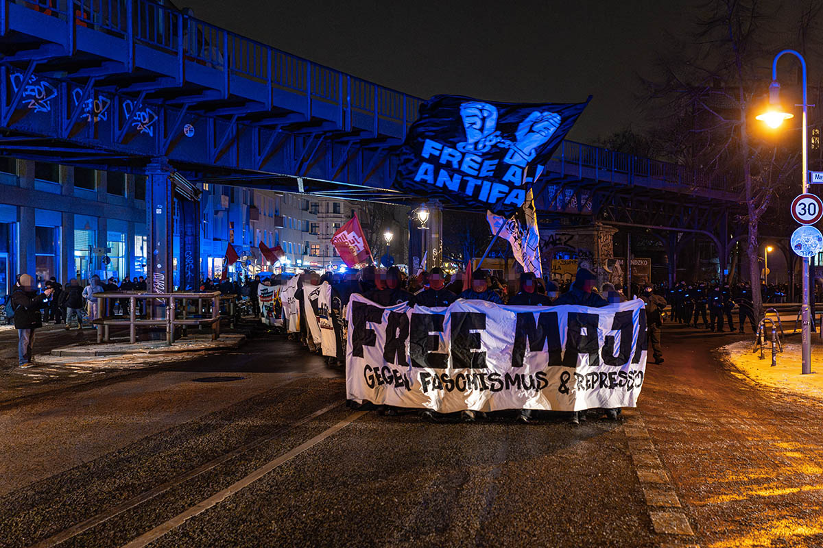 Das Foto zeigt die unter einer U-Bahn Brücke durchquerende Demo mit dem Fronttransparent mit dem Text "Free Maja - gegen Faschismus & Repression" darüber eine Fahne mit dem Text "Free all Antifas" und zwei aneinandergeketten Handen, die die Kette zerreißen.
