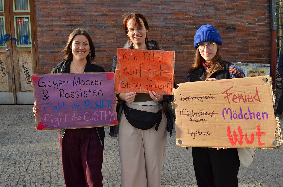 Das Foto von © Monika von Wegerer zeigt drei Teilnehmer*innen der Aktion mit selbstgemalten Plakaten: "Gegem Macker und Rassisten - Fight the Power - Fight the Cistem" "Kein Täter darf sich sicher fühlen" sowie Durchgestrichen: "Beziehungstat - Femizid" Durchgestrichen "Minderjährige Frauen - Mädchen" Durchgestrichen: "Hysterie - Wut"
