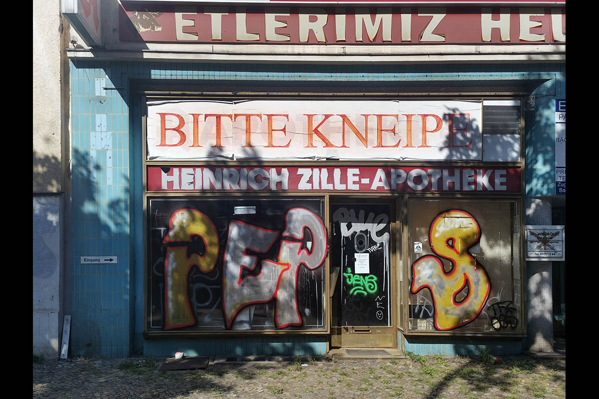 Das Foto zeigt ein verlassenes Ladengeschäft, über dessen Eingang ein Transparent mit dem Text "Bitte Kneipe" hängt. Darunter der frühere Schriftzug "Heinrich Zille-Apotheke". Einige Graffititags verzieren die Schaufensterscheiben