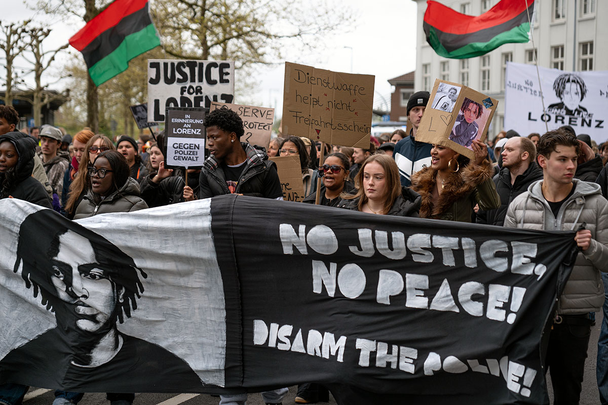 Das Foto von M. Goldschmidt zeigt einen Ausschnitt der vorderen Reihe der Demo mit Fronttransparenten - eines mit einem Bild von Lorenz A. dem Text "No Justice - no Peace - Disarm the Police"