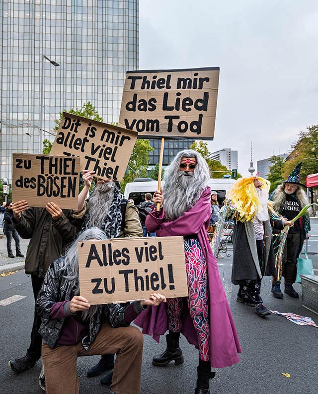 Das hochformatige Farbfoto zeigt eine Gruppe fantasievoll verkleideter Menschen bei einer satirischen Demonstration auf einer Berliner Straße. Mehrere Personen tragen lange, graue Perücken und künstliche Bärte, die an den Philosophen Friedrich Nietzsche erinnern. Sie halten handbeschriftete Schilder aus brauner Pappe mit Wortspielen über den Namen des Unternehmers Lars Windhorst beziehungsweise den früheren Karstadt-Eigentümer René Benko nicht – vielmehr beziehen sich die Sprüche auf den Namen „Thiel“. Im Mittelpunkt steht eine Person mit langem grauem Haar, einem ausladenden grauen Bart und einer kleinen bunten Sonnenbrille. Sie trägt einen auffälligen pinkfarbenen Mantel, darunter eine bunt gemusterte Hose und schwarze Stiefel. In der Hand hält sie ein großes Schild mit der Aufschrift „Thiel mir das Lied vom Tod“. Direkt davor kniet eine weitere verkleidete Person auf einem Bein. Sie trägt eine graue Langhaarperücke, eine dunkle Jacke und eine braune Hose. Vor ihrem Oberkörper hält sie ein Schild mit der Aufschrift „Alles viel zu Thiel!“. Links stehen weitere Demonstrierende mit Schildern, auf denen „Dit is mir allet viel zu Thiel“ und „zu THIEL des BÖSEN“ zu lesen ist. Rechts sind mehrere bunt kostümierte Personen zu sehen, darunter eine mit einer langen gelben Perücke und eine weitere mit einem spitzen Hexenhut. Einige tragen glitzernde Bänder und andere dekorative Accessoires. Im Hintergrund erhebt sich links ein großes, verglastes Hochhaus. Weiter entfernt ist der Berliner Fernsehturm zu erkennen. Fahrzeuge, Bäume mit herbstlich gefärbtem Laub und weitere Menschen säumen die Straße. Der Himmel ist hellgrau und bewölkt. Die Aufnahme wirkt humorvoll und theatralisch. Die bewusst übertriebenen Kostüme und die Wortspiele auf den Pappschildern verleihen der Demonstration den Charakter einer politischen Satire.