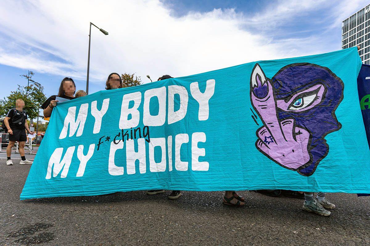 Das verpixelte Foto von © Björn Obmann zeigt das Transparent mit dem Text "My Body My (F*ching) Choice" neben einer maskierten Person, die den Stinkefinger zeigt.