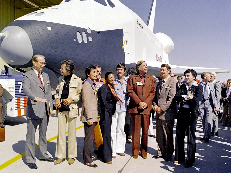 Das NASA-Shuttle Enterprise vor der Werksanlage in Palmdale mit der Star-Trek-Besetzung. Von links nach rechts: Dr. James C. Fletcher (damaliger Leiter der NASA), DeForest Kelley (Dr. „Pille“ McCoy), George Takei (Mr. Sulu), James Doohan (Chefingenieur „Scotty“ Scott), Nichelle Nichols (Lt. Uhura), Leonard Nimoy (Mr. Spock), Gene Roddenberry (der Erfinder von Star Trek), Kongressabgeordneter Don Fuqua und Walter Koenig (Fähnrich Pavel Chekov).