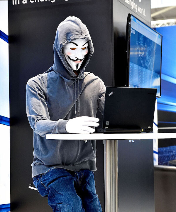 Foto: Frank Schwichtenberg / CC BY 4.0 Typische Darstellung eines Anonymous-Hackers mit Guy-Fawkes-Maske gekleidet in Jeans und Hoddie mit aufgesetzter Kapuze, der an einem Stehtisch sitzt und in eine Notebook tippt auf der Cebit 2016.