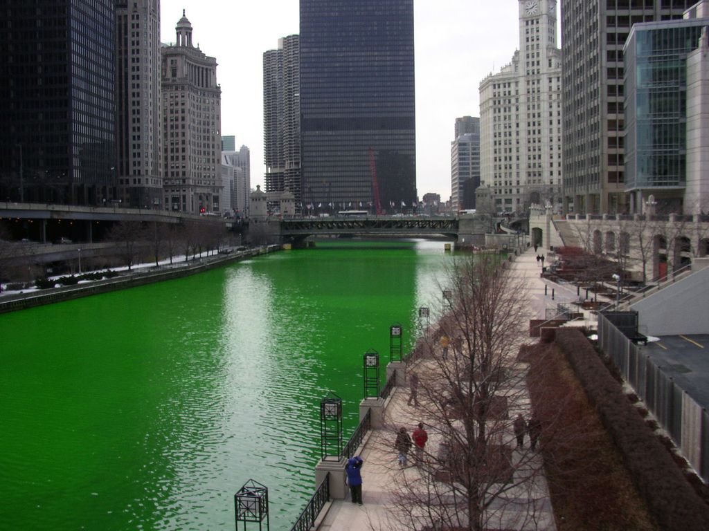 Das Foto zeigt den anlässlich des Saint Patrick’s Day traditionell grün gefärbten Chicago River an dessen Promenade Menschen spazieren und der von Wolkenkratzern umgeben ist.