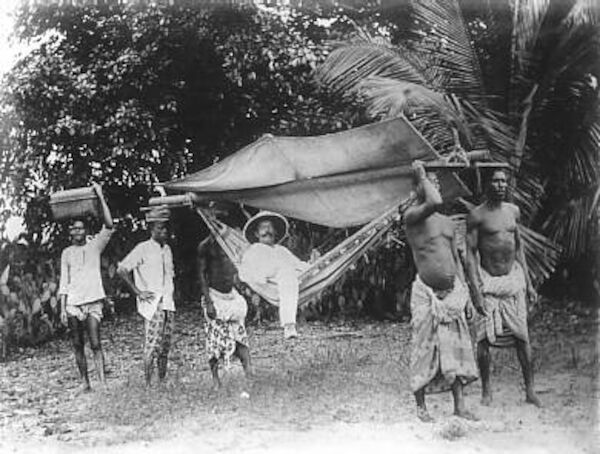 Das Foto zeigt einen deutschen Kolonialherren ca. im Jahr 1885, der sich von mehreren Trägern in einer Hängematte in Togo durch die Gegend tragen lässt.