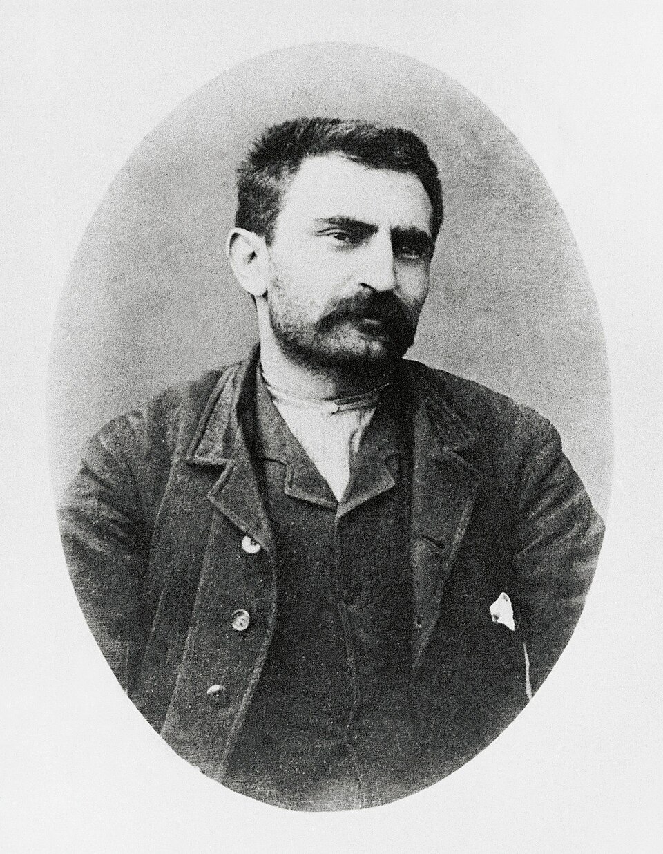 Historische Schwarz-Weiß-Fotografie des italienischen Anarchisten Errico Malatesta aus dem Jahr 1891, während seiner Zeit in der Illegalität. Das ovale Porträt zeigt ihn als Mann mittleren Alters mit kurzem, dunklem Haar, kräftigem Schnurrbart und dichtem Vollbart. Er trägt eine dunkle, leicht abgetragene Jacke über einer Weste und einem hellen Hemd. Seine Arme hält er hinter dem Rücken, den Kopf leicht zur Seite gedreht. Sein Gesichtsausdruck wirkt ernst und nachdenklich.
