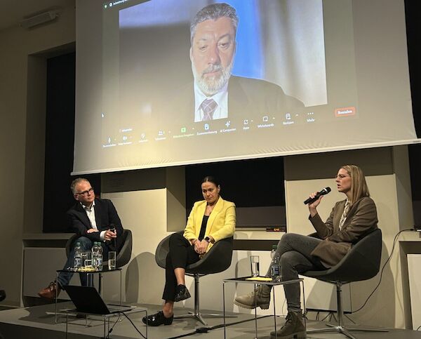 Das Bild zeigt eine Podiumsdiskussion mit 3 Personen, im Hintergrund eine Beamerleindwand