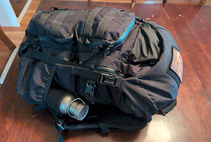 Das Foto zeigt einen liegenden, mit Molle System ausgerüsteten schwarzen Rucksack mit einer Edelstahltrinkflasche in der rechten Seitentasche