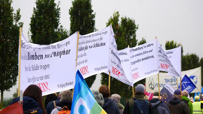 Fotografie einer Demonstration im Freien. Von hinten ist eine Gruppe von Menschen in Jacken zu sehen, die zahlreiche Fahnen und große weiße Transparente mit schwarzen und roten Schriftzügen tragen. Auf den Transparenten stehen Zitate von Wolfgang Borchert, darunter „Dann gibt es nur eins: SAG NEIN!“. Dazwischen wehen blaue Friedensfahnen, eine davon mit einer weißen Friedenstaube. Im Hintergrund stehen hohe grüne Bäume unter einem grauen, bewölkten Himmel.
