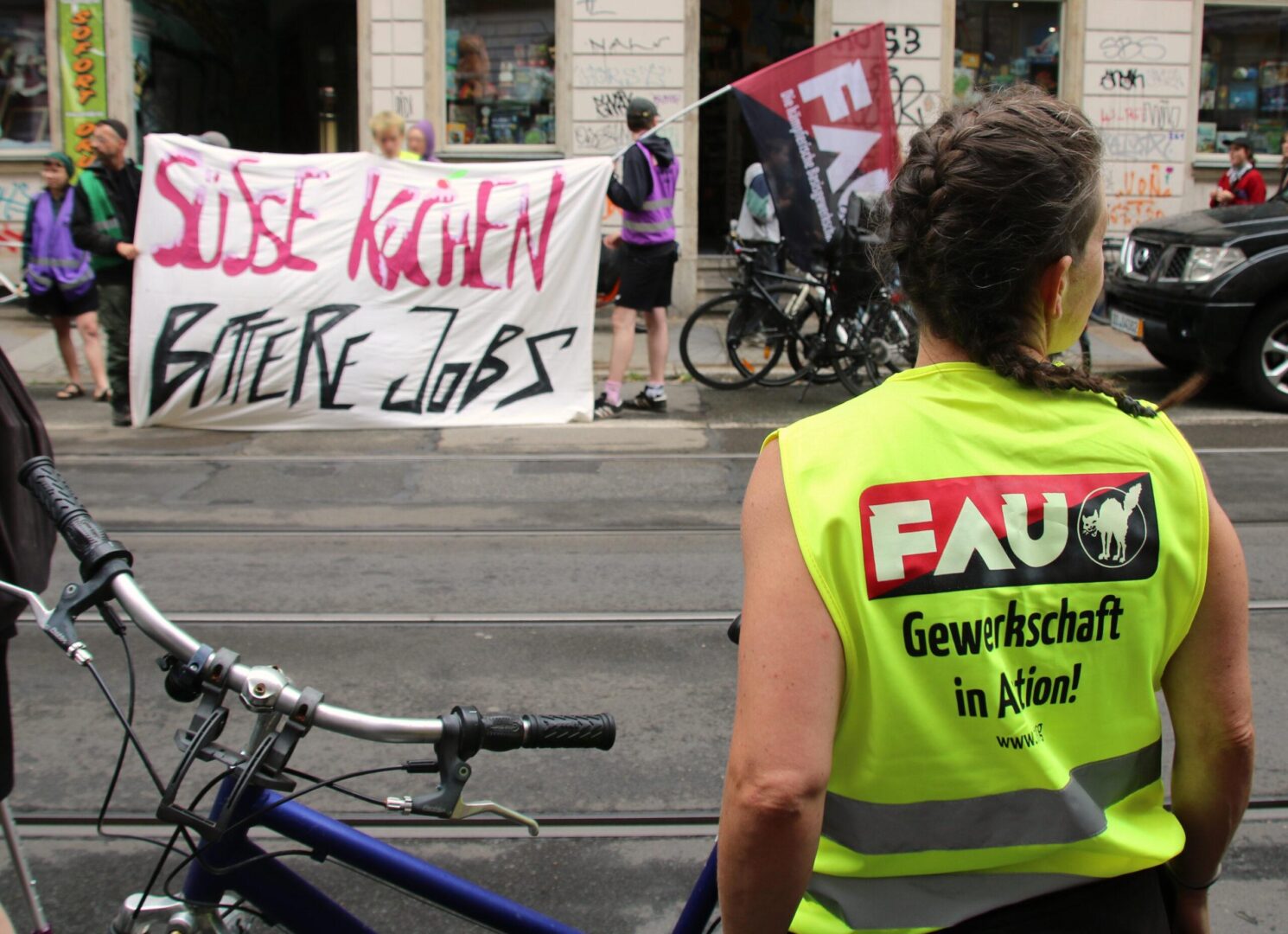 Aktivist:Innen mit Transparent "Süße Kuchen - bittere Jobs". Eine Person im Vordergrund mit "FAU - Gewerkschaft in Aktion" Weste
