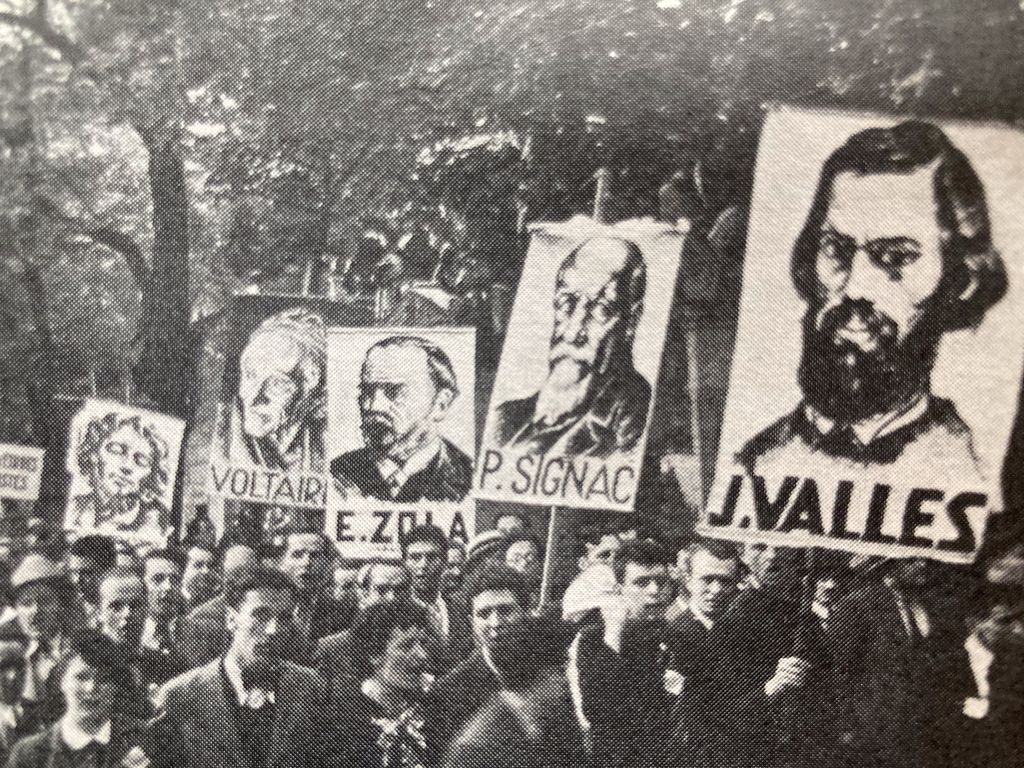 Das historische Foto zeigt die Menschenmassen vor dem Friedhof mit den Portraits verschiedener Persönlichkeiten der französischen Arbeiter- und Volksbewegung wie Voltaire, Emile Zola, Jules Vallès, Paul Signac