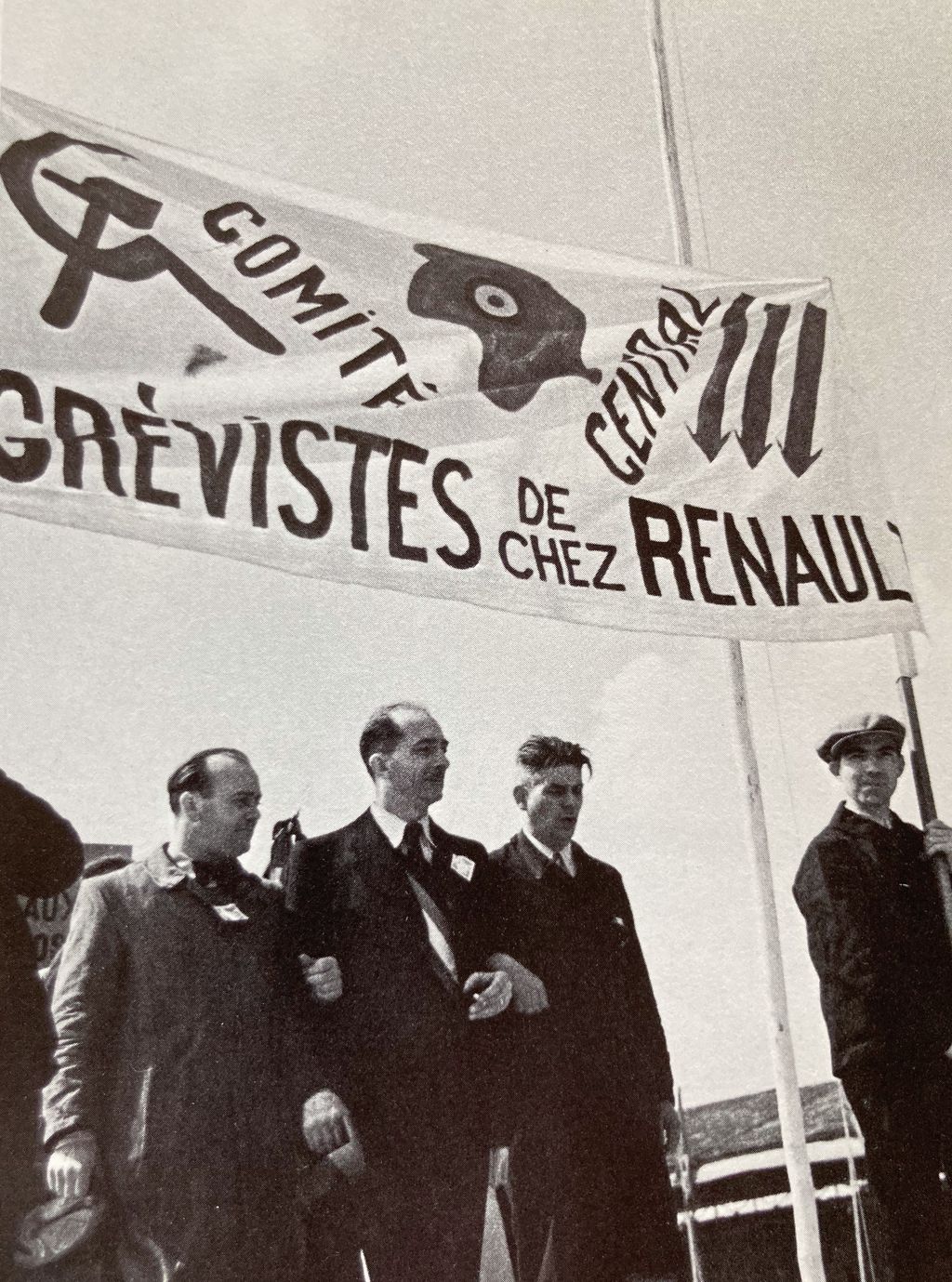 Das historische Foto zeigt Aktivisten des Streiks unter einem Banner des Zentralkomitees mit Hammer und Sichel, der Kommunardenkappe und der 3 nach unten gerichteten Pfeile, ähnlich derjenigen der Eisernen Front in Deutschland Anfang der 1930er Jahre