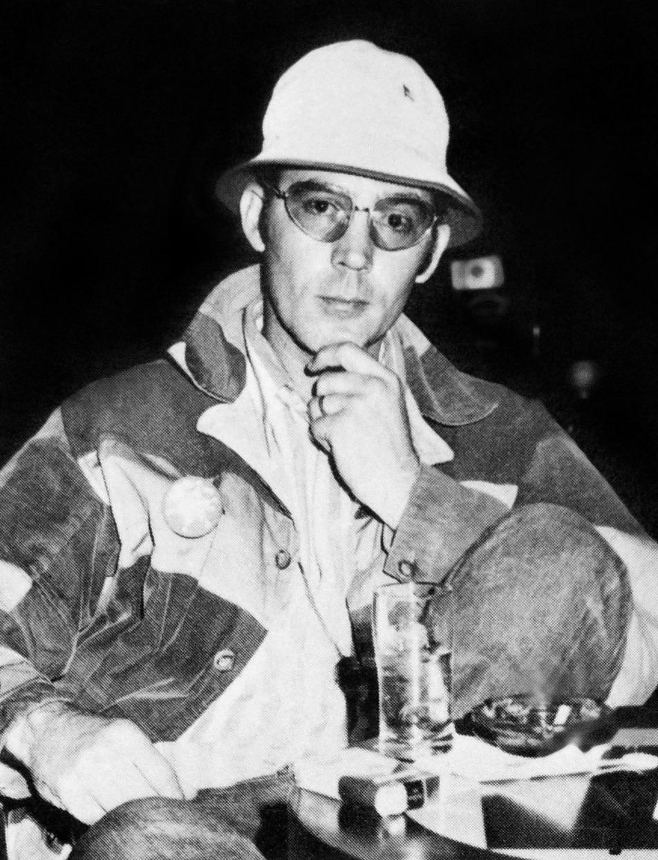 A black and white photo of Hunter S. Thompson wearing glasses, a white hat, and a patterned jacket. He is sitting in a chair with one hand on his chin, appearing thoughtful. A glass of drink and a small dish are on the table in front.