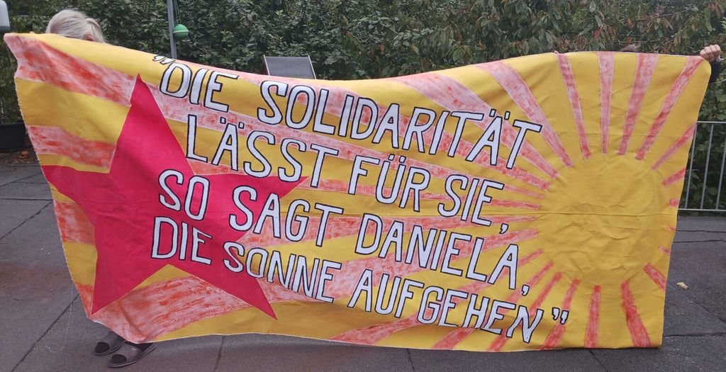Das Foto zeigt ein von mehreren nicht oder nur teilweise sichtbaren Personen getragenes Transparent mit dem Text: "Die Solidarität lässt für sie so sagt Daniela die Sonne aufgehen". Links vom Text ein roter Stern, rechts davon eine Sonne.