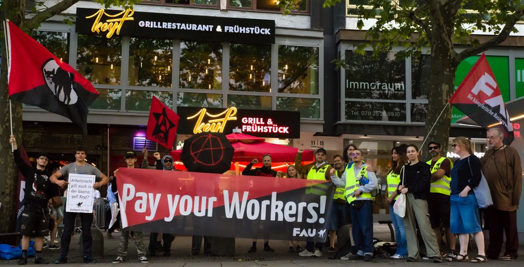 Das Foto zeigt die Solikundgebung vor dem Keyf am 17. Juli 2026 mit solidarischen Teilnemer*innen und dem "Pay Your Workers" Transpartent sowie einige FAU Fahnen und weitere, unter anderem mit dem Anarcho A