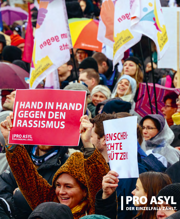 Das Foto zeigt eine Demo mit einer Frau, die eine Pro Asyl Tafel mit dem Text "Hand in Hand gegen Rassismus" hoch hält.