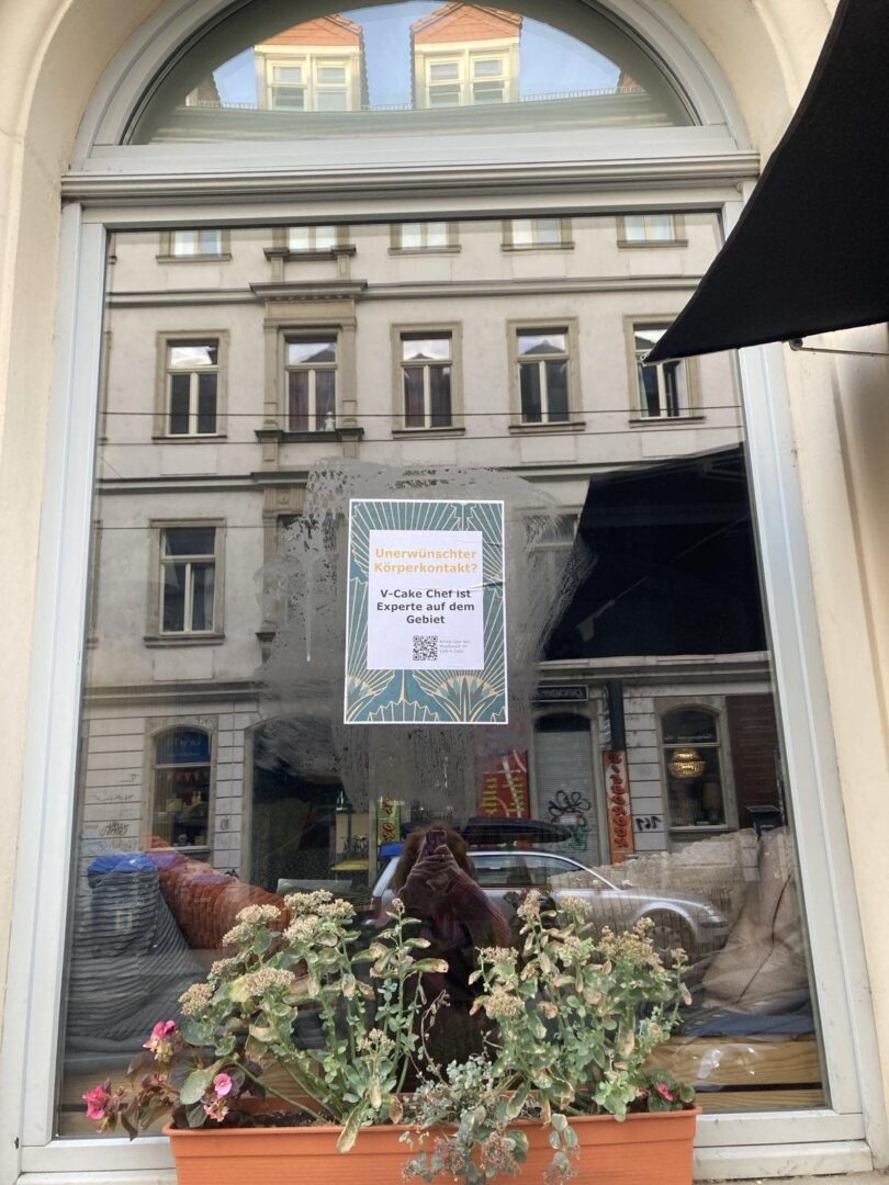 An einer Fensterscheibe klebt ein Plakat mit dem Text "unerwünschter Körperkontakt? V-Cake Chef ist Experte auf dem Gebiet"